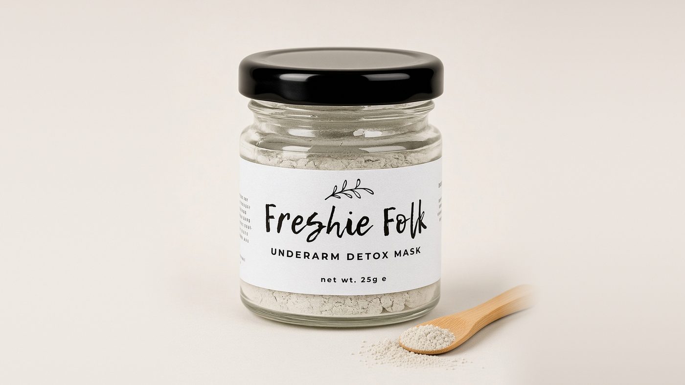 A small glass jar of Freshie Folk Underarm Detox Mask clay powder with a black lid, beside a bamboo spoon of pale clay on a soft cream background.