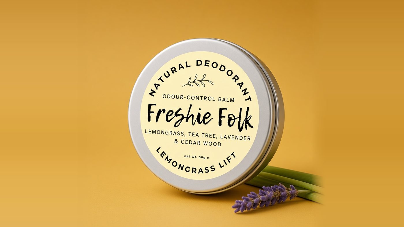A Freshie Folk Lemongrass Lift deodorant balm tin with a pale yellow label on a warm mustard-yellow background, with lemongrass stems and a sprig of lavender beside it.