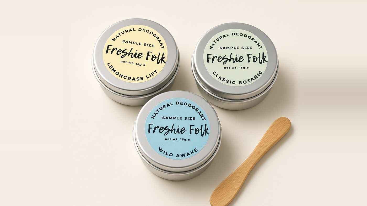 Three Freshie Folk sample-size deodorant balm tins, Lemongrass Lift with a yellow label, Classic Botanic with a green label and Wild Awake with a blue label, beside a wooden bamboo applicator on a pale cream surface.